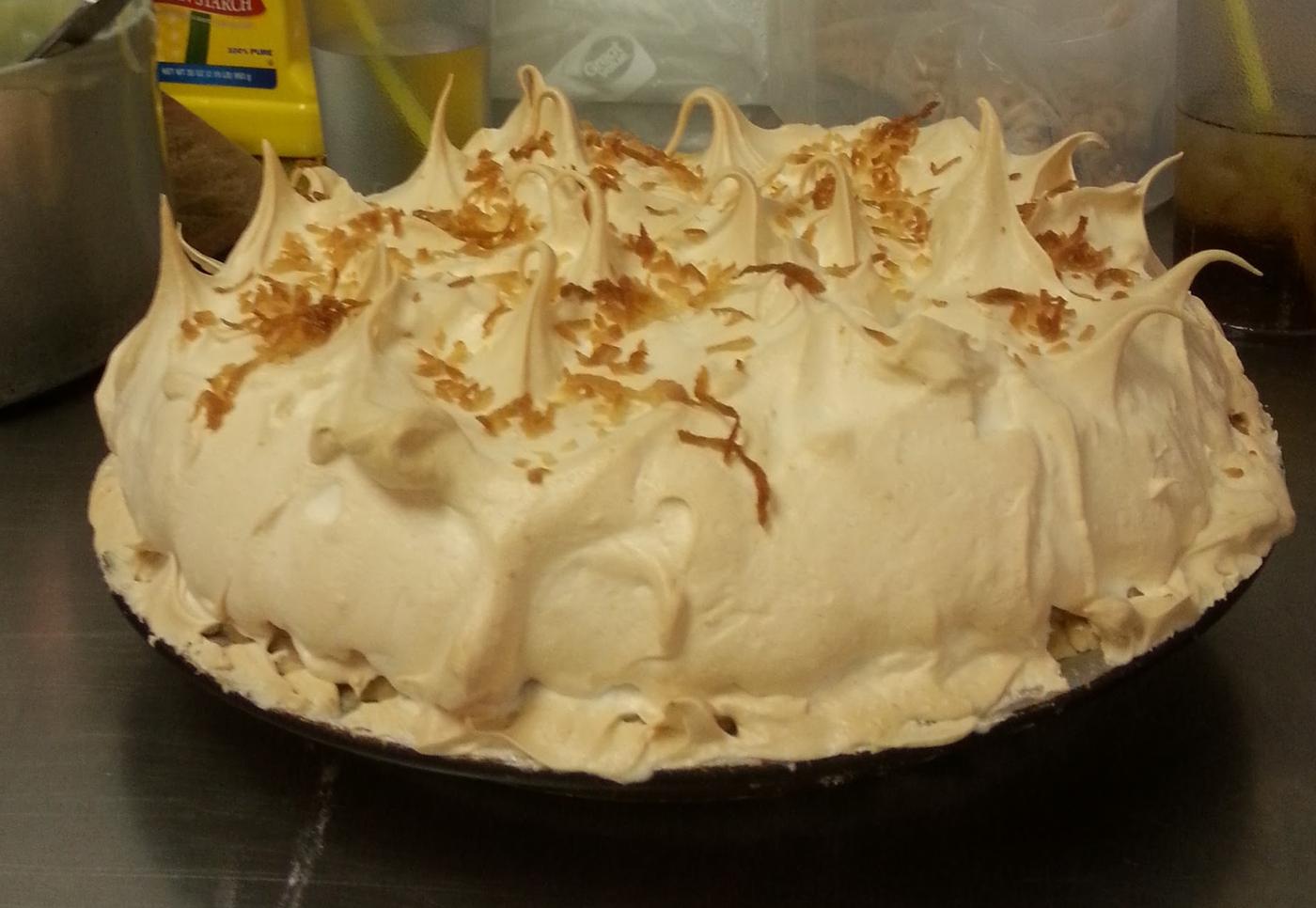 A coconut pie under a tall torched meringue, on the kitchen bench